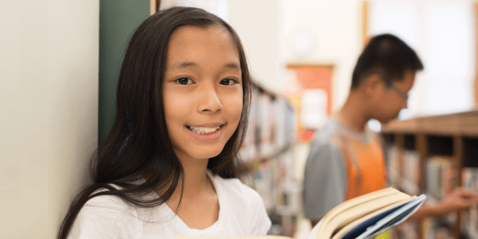 A young girl holds a book in a library; in the background, another patron looks at a book on a shelf.