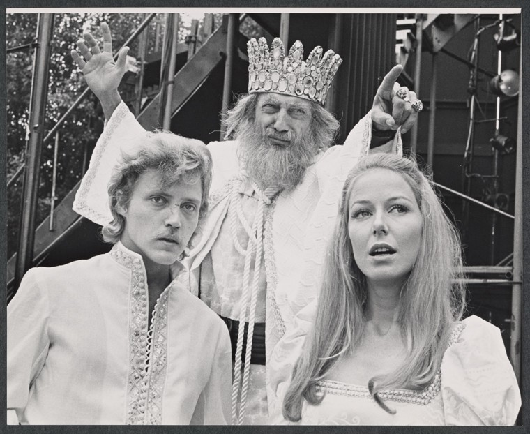 man and woman dressed in white with older man behind them wearing crown and pointing