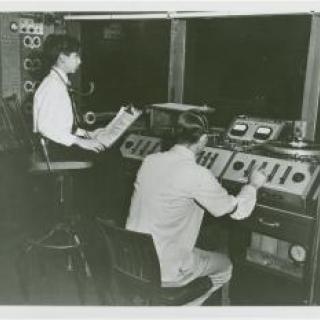 Black and white photo of two people at soundboard.