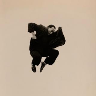 A Black man in mid-air with his knees pulled toward his chest, arms bent, looking down and wearing black garments and shoes