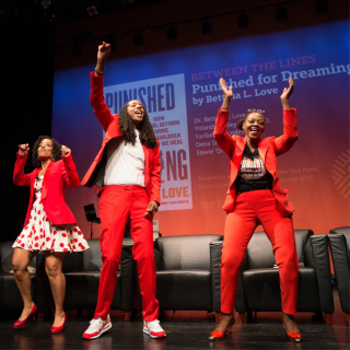 Three people standing onstage at the Schomburg Center. They are standing up and waiving their hands in the air, appearing to applaud.