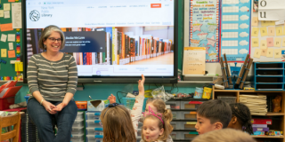 An educator sits in front of a large screen displaying NYPL's homepage and smiles to a classroom full of engaged, young students