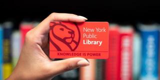 A hand holding an NYPL card in front of a row of books